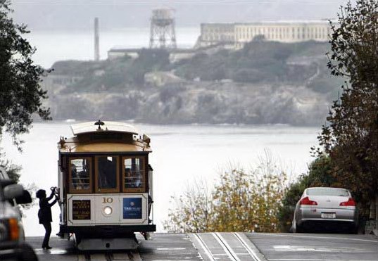 An emission-free San Francisco cable car makes a stop on Russian Hill, with Alcatraz Island in the background.